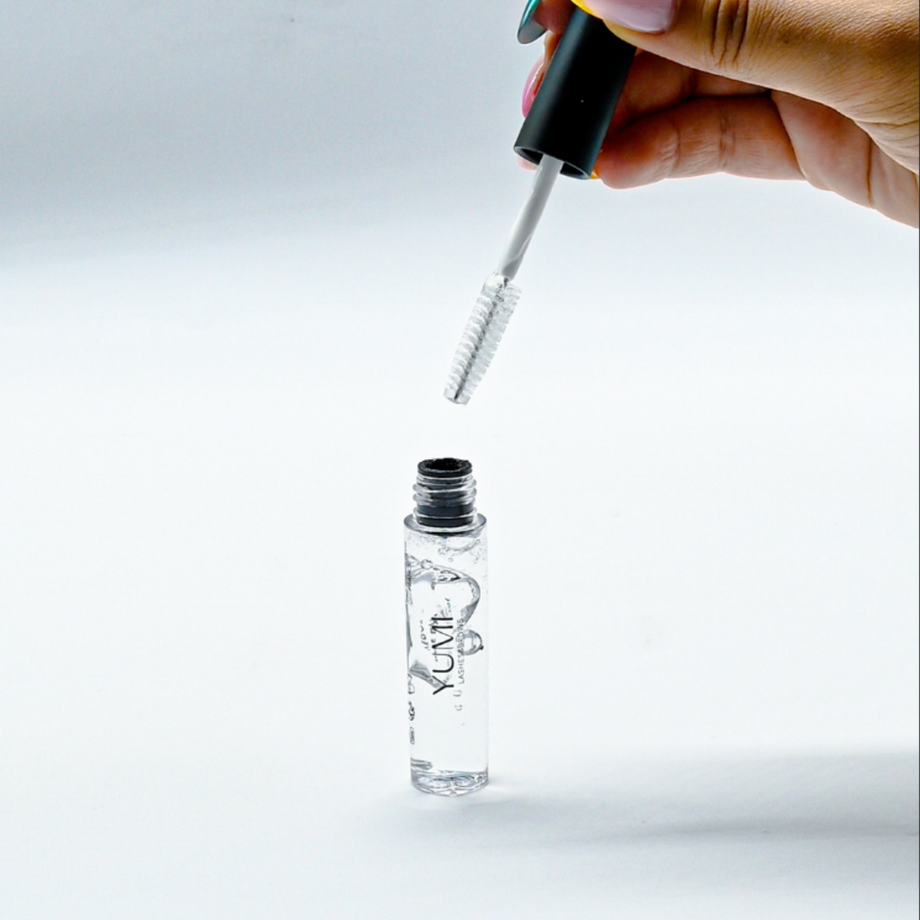 Eyelash serum with applicator held above a clear bottle labeled 'YUMI' on a light gray background