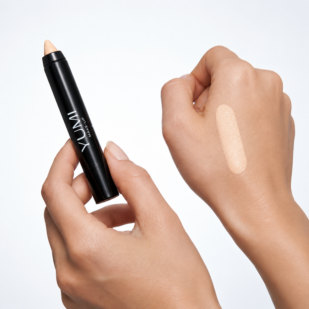 A black Yumii makeup stick with a light beige cream product, held by a hand next to a hand swatch showing the cream's texture and shade.
