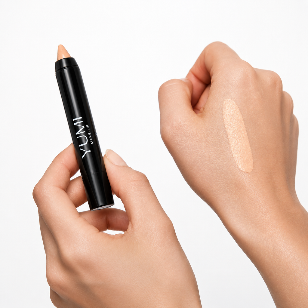 A YUMI makeup concealer stick held in a hand, with a swatch of the beige concealer applied on the back of another hand, against a clean white background.